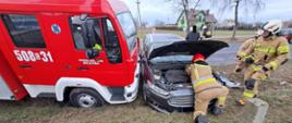 Zdjęcie przedstawia samochód osobowy oraz wóz strażacki po kolizji drogowej, strażaków PSP i OSP podczas prowadzenia działań.