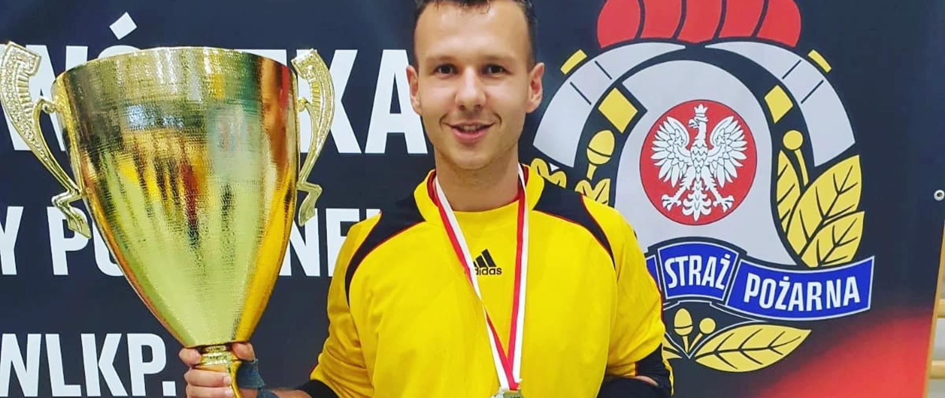 Na zdjęciu widać mężczyznę w stroju sportowym, spodenki koloru białego koszulka koloru żółtego. W ręku mężczyzny znajduje się złoty puchar. W tle widać logo Państwowej straży pożarnej. 