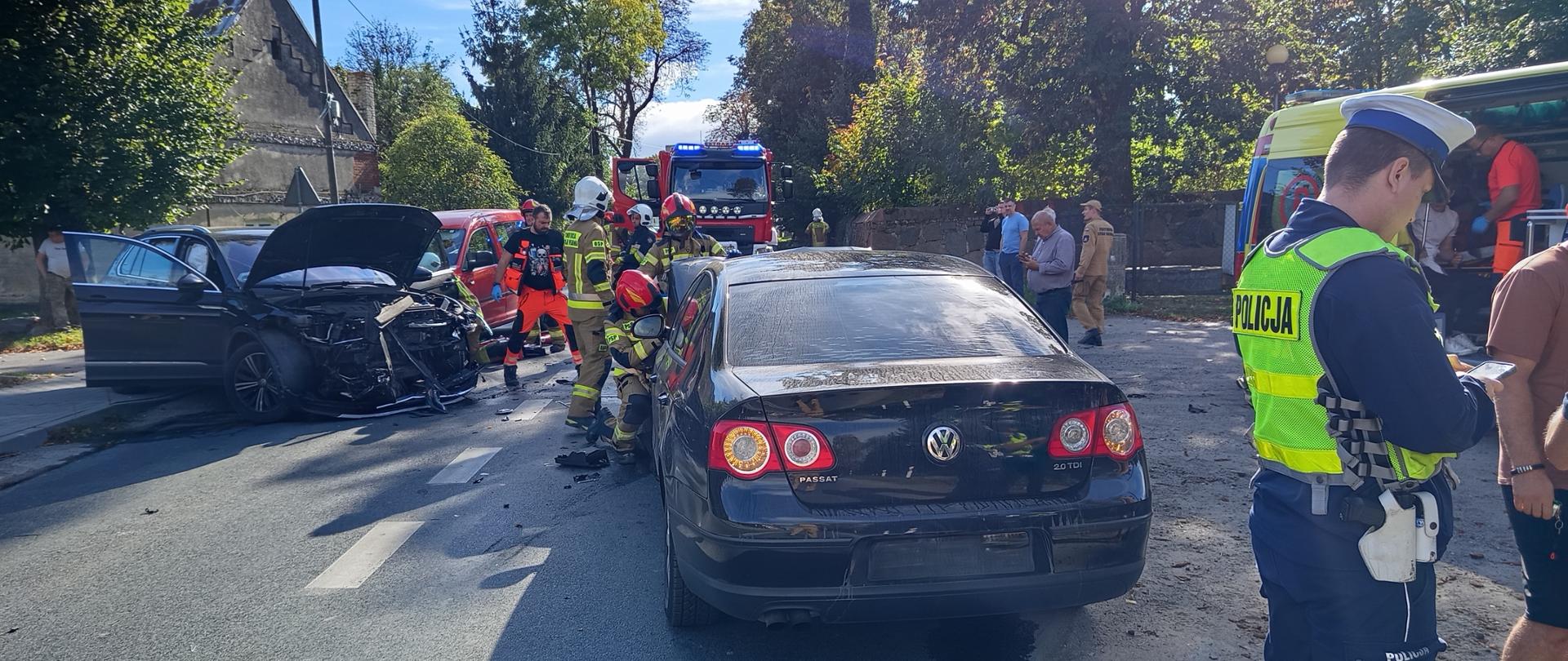 Fotografia przedstawia ujęcie wzdłuż drogi powiatowej, na pierwszym planie widoczny od tyłu pojazd osobowy marki Volkswagen passat, na dalszym planie widoczny inny pojazd stojący przodem do fotografującego, z rozbitym przodem, jeszcze dalej w tle widoczny pojazd pożarniczy, przy rozbitych pojazdach pracują strażacy i ratownicy medyczni.