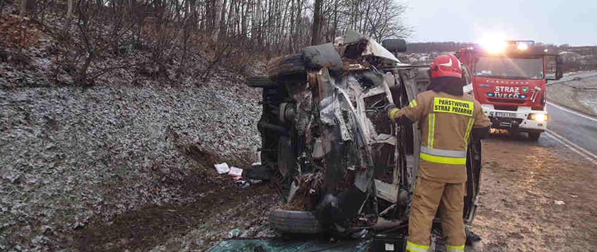 Zdjęcie przedstawia strażaka odłączającego akumulator w rozbitym samochodzie dostawczym. Na drugim planie samochód ratowniczo-gaśniczy stojący na jezdni