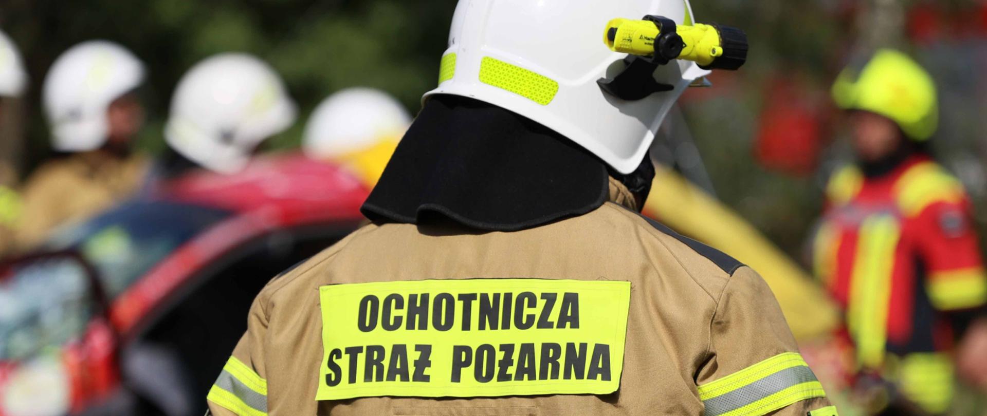 Mężczyzna w stroju bojowym straży pożarnej z napisem Ochotnicza Straż Pożarna zbliża się do grupy innych strażaków przewracających rozbity samochód.