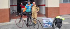 Na tle stojącego w garażu samochodu pożarniczego, przed budynkiem komendy stoi strażak w ubiorze kolarskim, obok zastępca komendanta powiatowego ubraniu służbowym w kolorze piaskowym. Mężczyźni obejmują sie za ramiona. Przed nimi stoi kolarzówka z doczepiona przyczepką jednokołową. 
