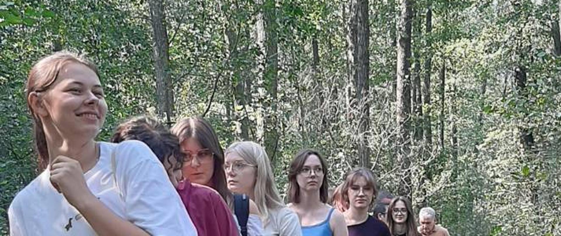 Grupa dziewczyn, na końcu mężczyzna, idzie po wąskiej drewnianej ścieżce przez las