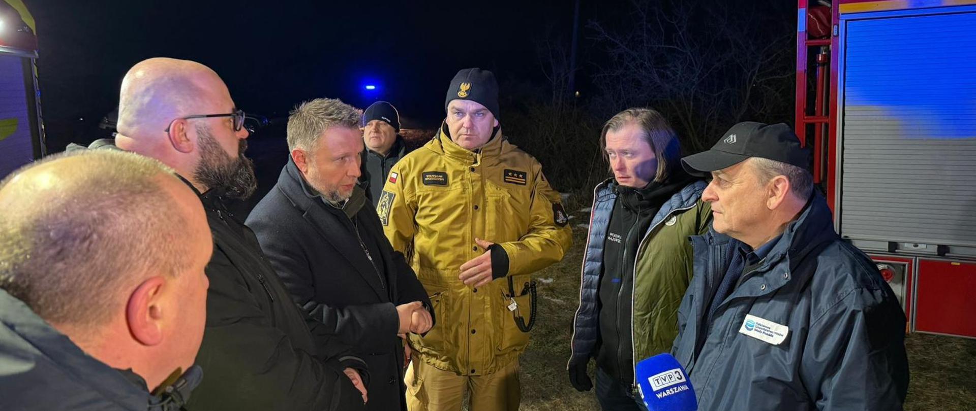 Grupa mężczyzn, w tym strażak w kurtce służbowej PSP, prowadzi rozmowę w nocy przy samochodzie pożarniczym. Jeden z obecnych udziela wypowiedzi do mikrofonu telewizyjnego. Spotkanie odbywa się w terenie podczas działań ratowniczych.