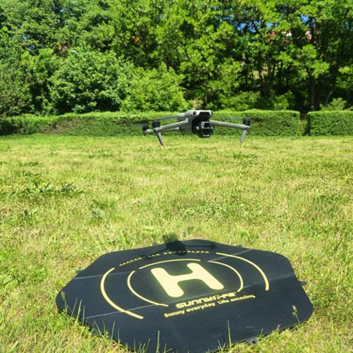 otografia przedstawia bezzałogowy statek powietrzny (dron) marki DJI, model Air 3S, sfotografowany od przodu podczas zawisu w powietrzu. Dron znajduje się około metra nad ziemią, bezpośrednio nad rozłożonym na zielonej trawie ośmiokątnym, czarnym lądowiskiem marki Sunnylife z żółtym oznaczeniem lądowania „H”. Tło zdjęcia to jasny, nasłoneczniony trawnik, za którym ciągnie się ściana gęstej, zielonej roślinności, krzewów i drzew. Światło jest dzienne, mocne, charakterystyczne dla bezchmurnego, letniego dnia.
