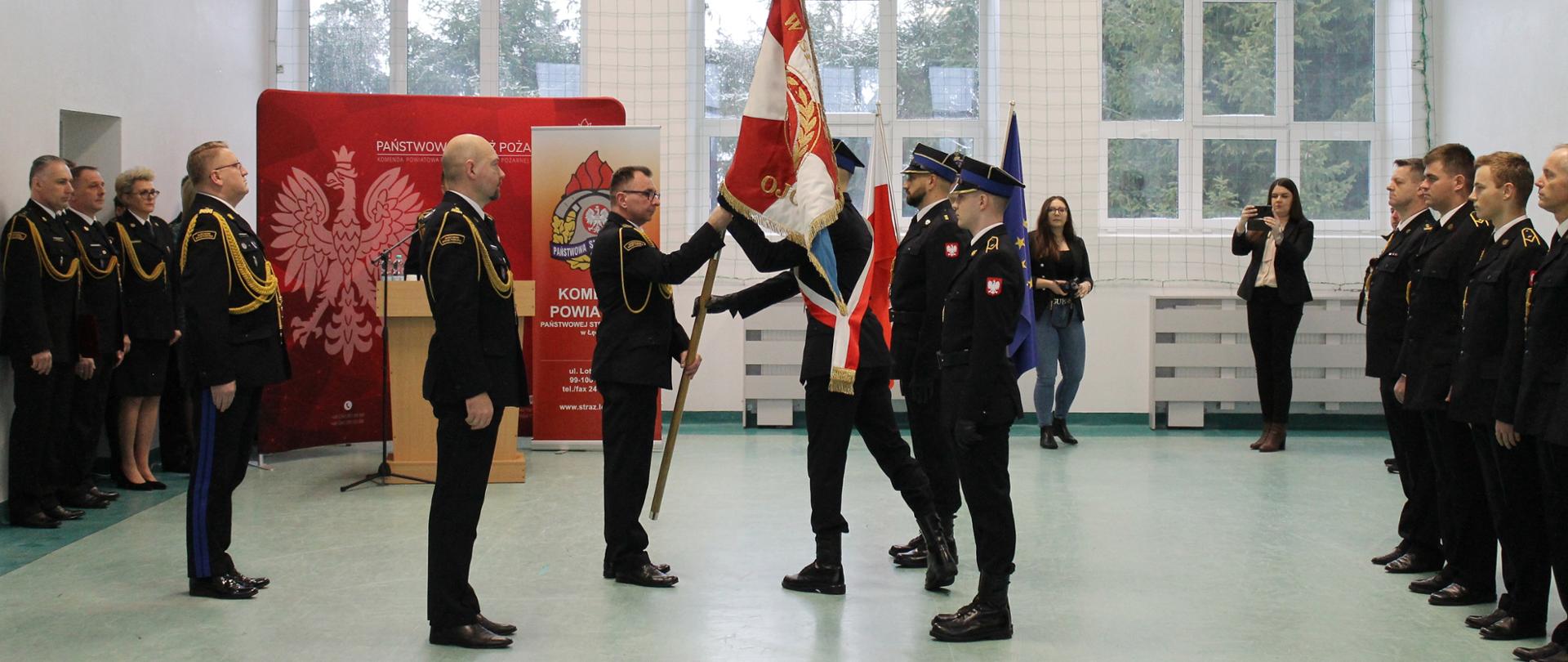 brygadier Piotr Jóźwiak wita się ze sztandarem komendy, w hali sportowej są jeszcze strażacy i przedstawiciele władzy rządowej i samorządowej