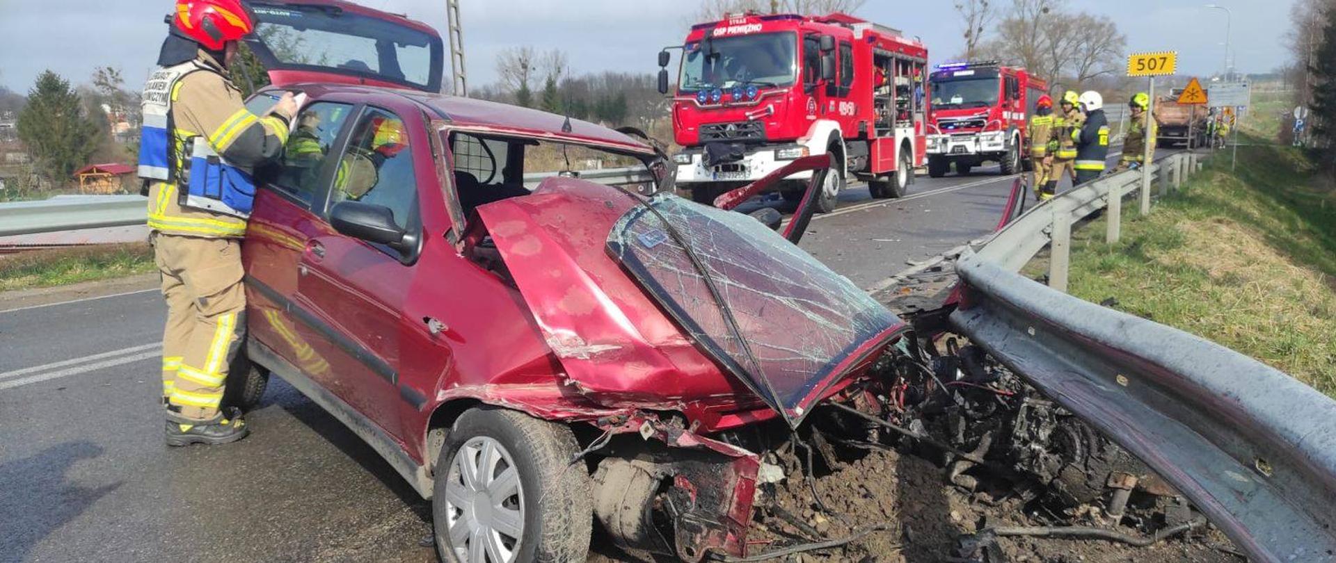 Na drodze w poprzek jednego pasa ruchu stoi czerwone uszkodzone po zderzeniu auto osobowe. Przy aucie strażak w jasnym mundurze i czerwonym hełmie. Dalej na drodze dwa czerwone auta strażackie. Słonecznie
