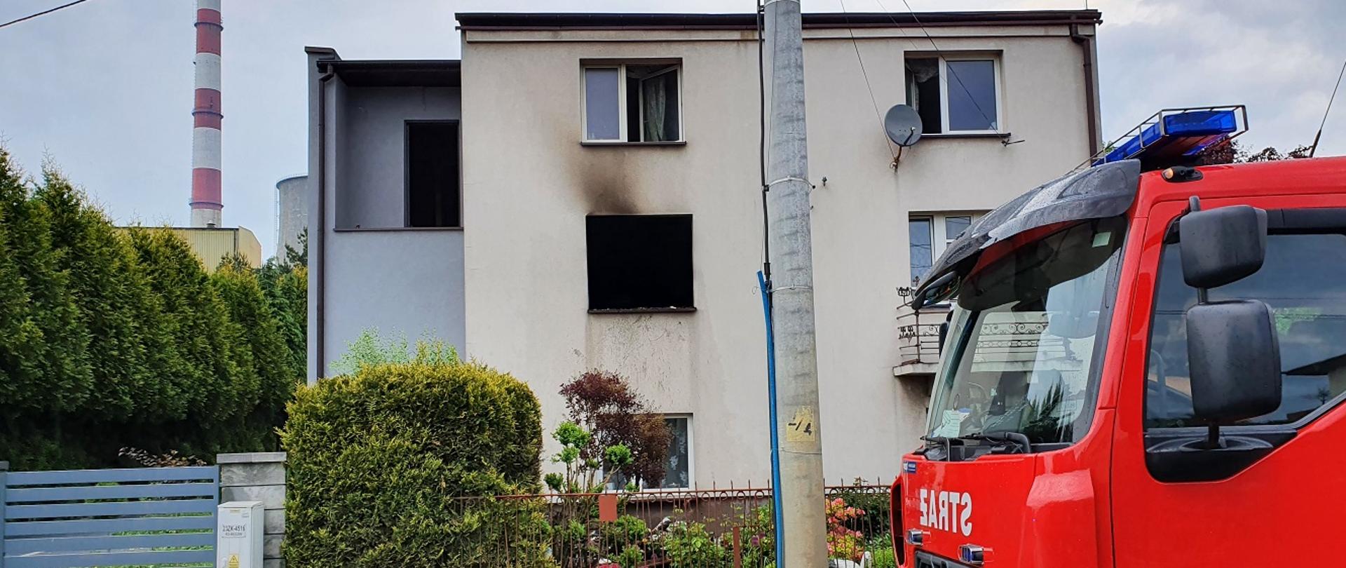 Na zdjęciu widać kabinę samochodu pożarniczego stojącego na jezdni. W tle widać budynek mieszkalny w którym miejsce miał pożar (budynek nie ma okna oraz ma okopconą ścianę).