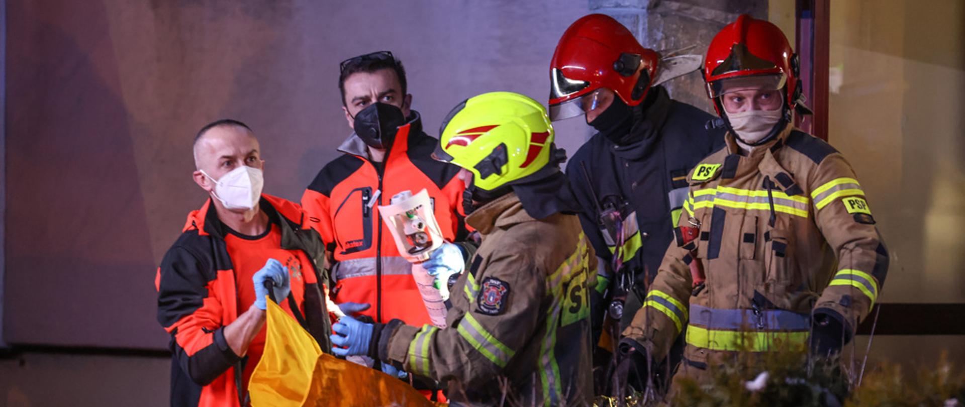 Strażacy i Ratownicy Medyczni ewakuujący osobę poszkodowaną z pożaru.