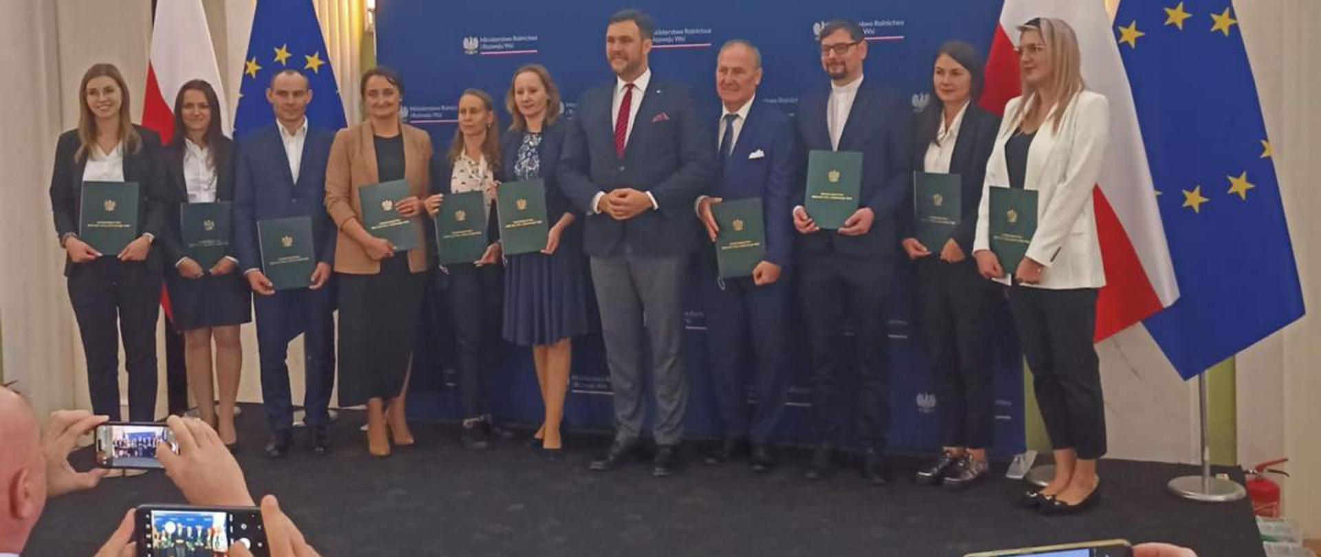 Pamiątkowe zdjęcie grupowe na podium. Grupa osób trzymających zielone teczki stoi obok oficjeli na tle ścianki z logo Ministerstwa Rolnictwa i Rozwoju Wsi. Na pierwszym planie widoczne dłonie osób fotografujących wydarzenie telefonami.