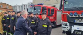 Mężczyzna w garniturze wita się ze strażakami za nimi stoją samochody gaśnicze.
