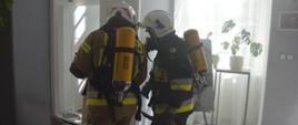 Ćwiczenia w Pakosławiu. Dwóch strażaków wyposażonych w aparaty oddechowe prowadzi działania wewnątrz budynku. Jeden z nich ma na sobie ubranie koloru piaskowego, drugi granatowego. Trzymają w rękach wąż i prądownicę. W tle okno i kwiatek.