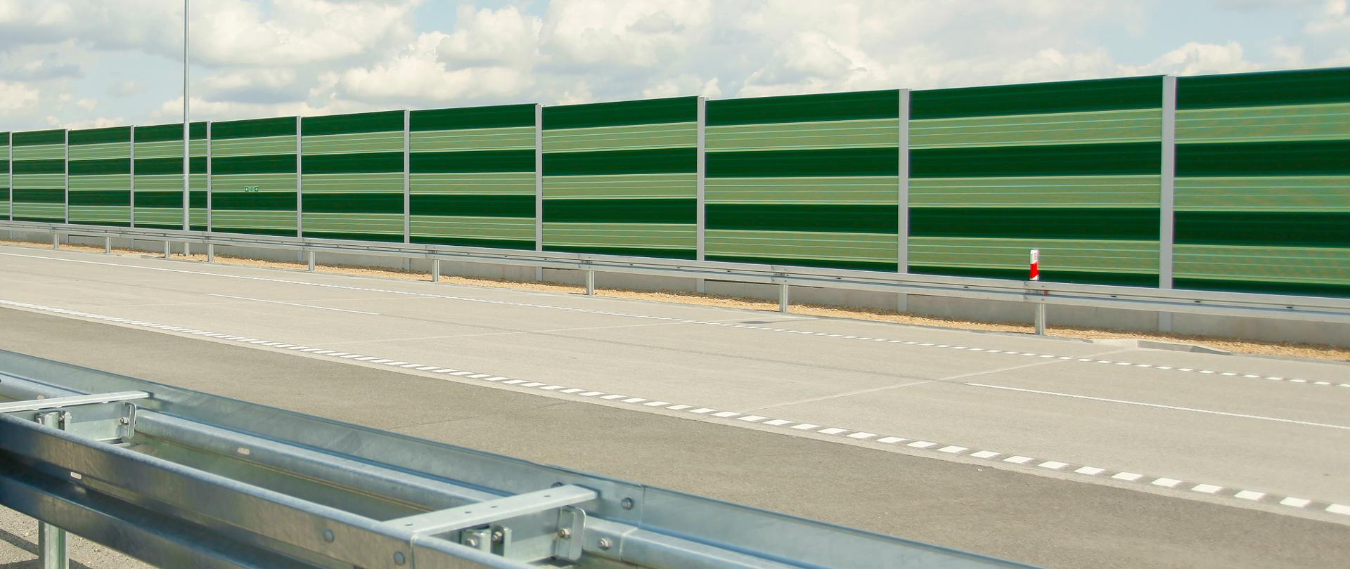 Obraz przedstawia fragment nowo wybudowanej lub dobrze utrzymanej drogi z widoczną barierą ochronną z metalu na pierwszym planie oraz zieloną barierą dźwiękochłonną w tle. Na drodze widać wyraźne oznaczenia pasów oraz gładką nawierzchnię, co świadczy o jej wysokiej jakości. Zielona bariera dźwiękochłonna, składająca się z paneli wspartych na metalowych słupach, ma na celu redukcję hałasu drogowego dla okolicznych terenów.