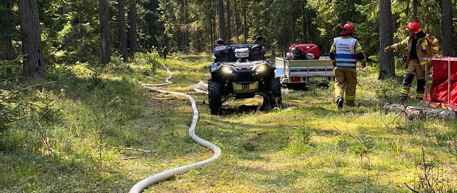 Na drodze leśnej rozciągnięty jest wąż strażacki. W oddali stoi quad i dwóch strażaków.