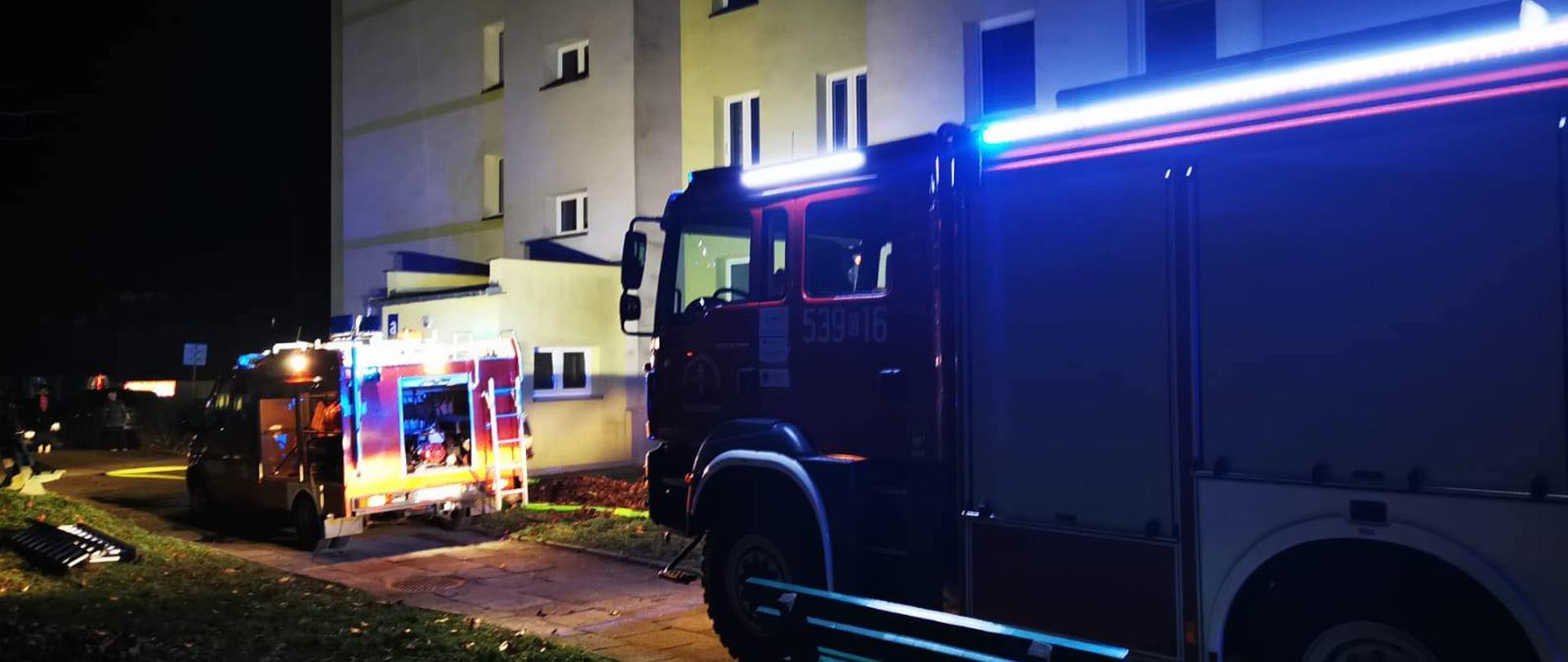 pora nocna. przed budynkiem oświetlonym przez światła samochodów stoją dwa pojazdy pożarnicze.