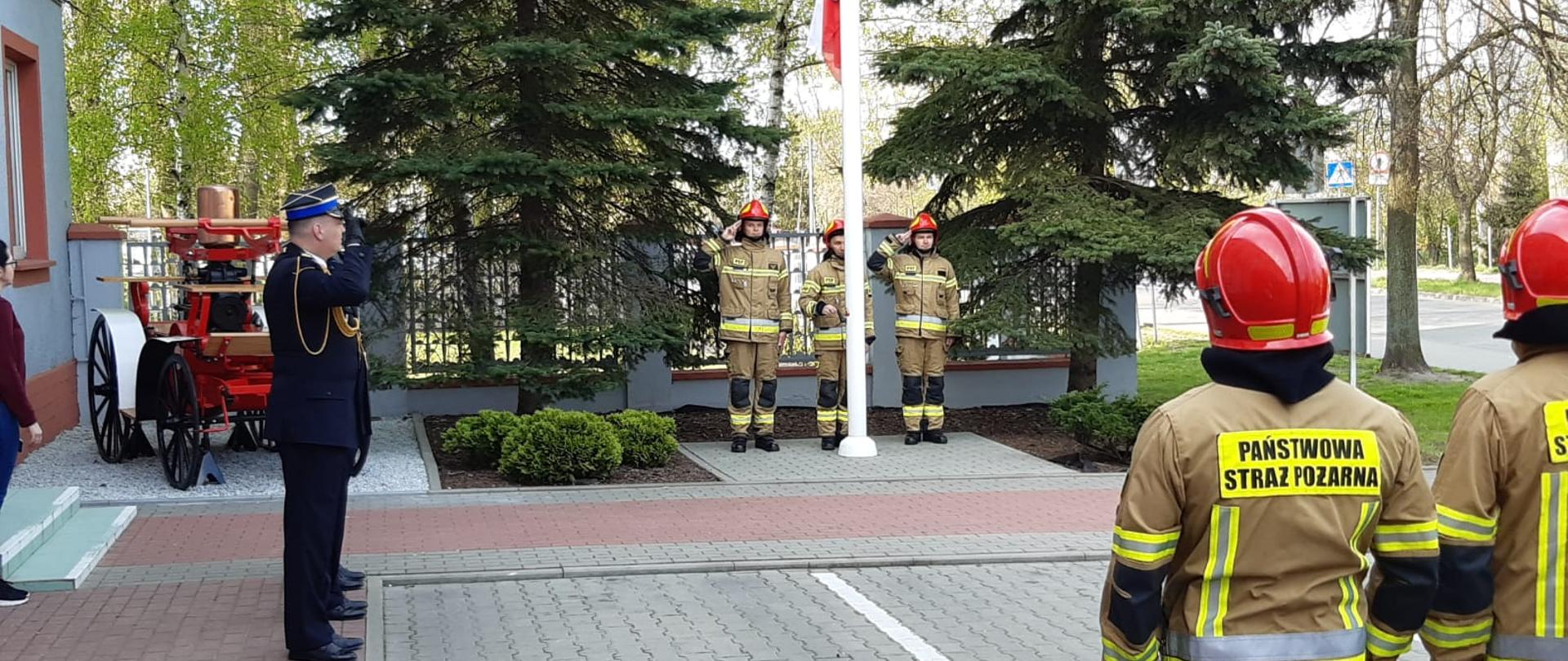za zdjęciu strażacy stojący na zbiórce 1 ma mundur wyjściowy reszta jest w ubraniach bojowych i czerwonym hełmach, trzech stoi za masztem flagowym dwóch po prawej stronie, w tle dwa drzewa sikawka i ogrodzenie