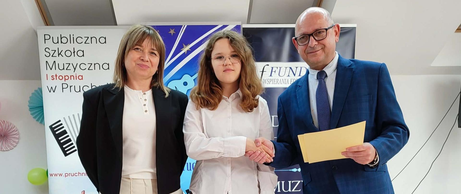 Na scenie Amelia Lewkowicz – II nagroda w kategorii III w czasie ogłoszenia wyników konkursy wraz z organizatorem Grzegorzem Wójcikiewiczem i nauczycielem Iwoną Grabowską. W tle znajdują się banery związane z wydarzeniem muzycznym oraz szkołą muzyczną w Pruchniku. 