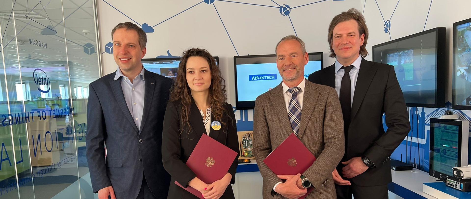 Cztery osoby - trzech mężczyzn w garniturach i kobieta stoją w małym pokoiku, za nimi monitory.