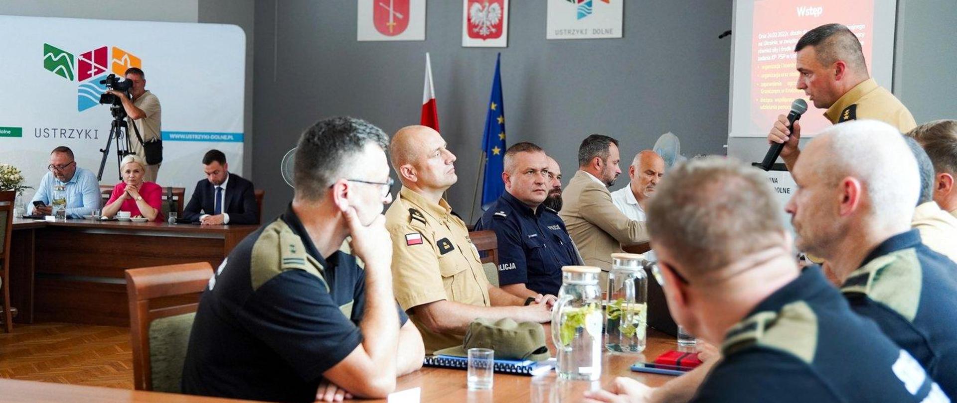 Wizyta posłów Sejmowej Komisji Administracji i Spraw Wewnętrznych w Ustrzykach Dolnych. Zdjęcie zrobione w sali konferencyjnej podczas narady. Przy stołach siedzą uczestnicy, wśród nich oficerowie Państwowej Straży Pożarnej, Policji, Straży Granicznej.