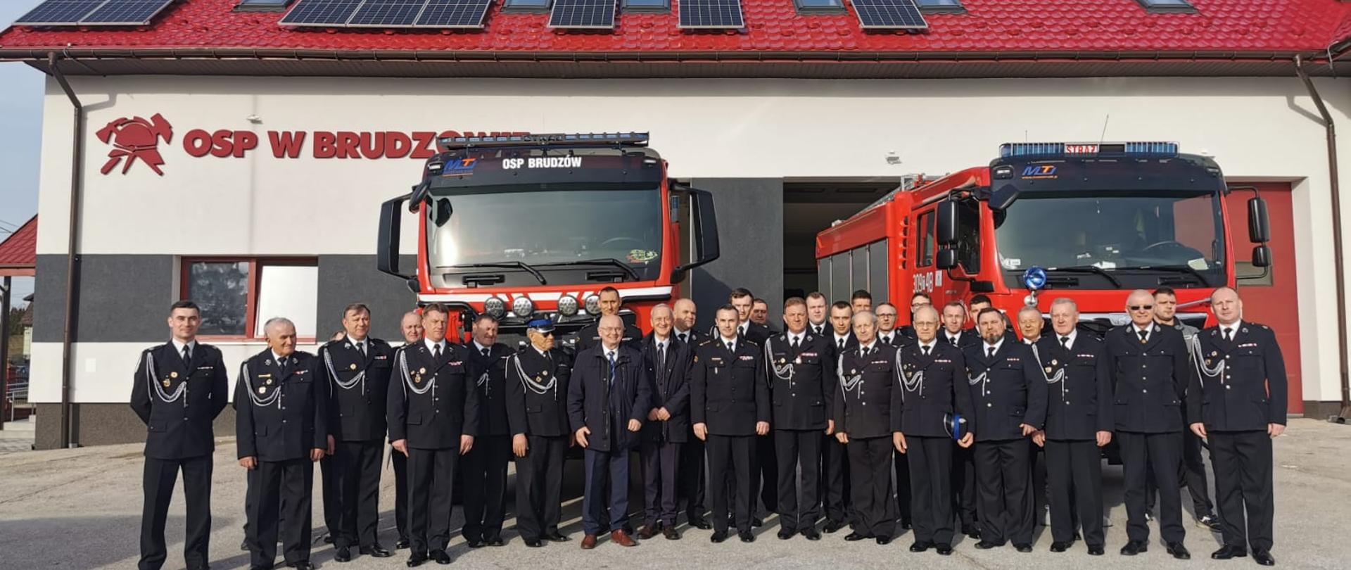 Zdjęcie przedstawia druhów, gości i organizatorów zebrania sprawozdawczego Ochotniczej Straży Pożarnej w Brudzowie. Na tle samochodów pożarniczych i budynku remizy.