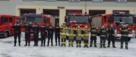 Strażacy z Komendy Powiatowej Państwowej Straży Pożarnej w Kozienicach tworząc z dłoni serca stoją na tle budynku komendy i samochodów pożarniczych