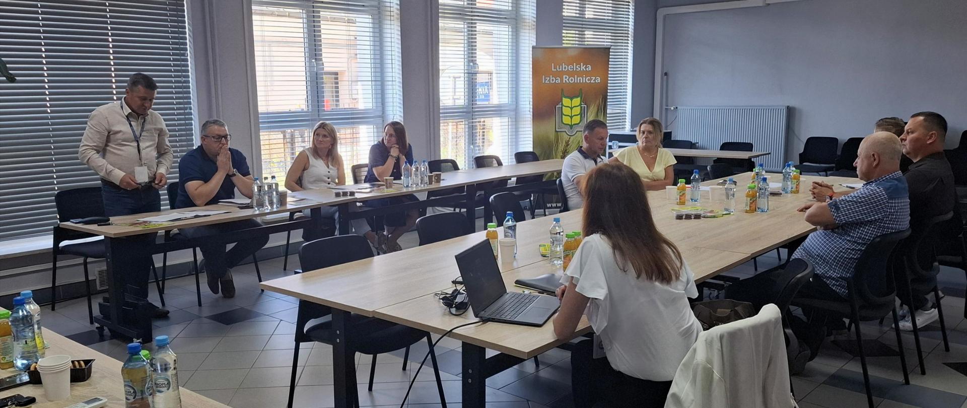 Kilka osób siedzi przy stołach na sali konferencyjnej. Jeden mężczyzna stoi i przemawia.