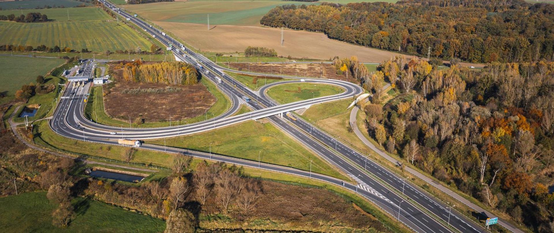 Widok z lotu ptaka na węzeł drogowy z estakadą nad autostradą. Wokół węzła znajdują się pola uprawne, tereny zielone oraz las o jesiennych barwach. Na drogach widoczne są pojazdy poruszające się po autostradzie i łącznicach.