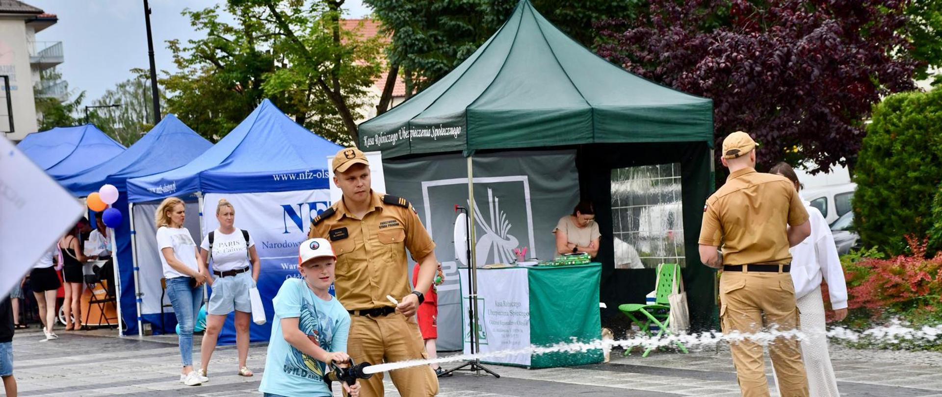 Dziecko ćwiczy gaszenie pożaru wodą pod nadzorem strażaka.