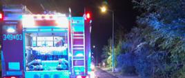 Na zdjęciu wykonanym w porze nocnej widać pojazd straży pożarnej w kolorze czerwonym. Zdjęcie zrobione przed budynkiem w którym powstał pożar. Teren oświetlany jest sztucznym światłem. Widać ulice i po bokach zielone zarośla.