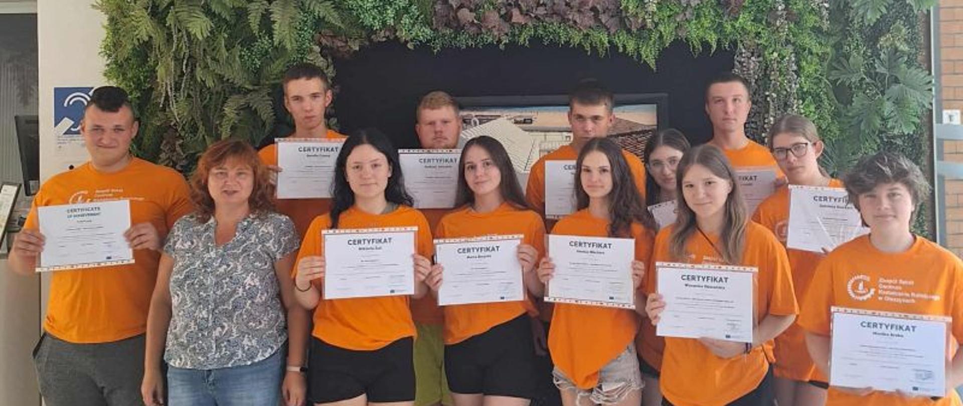Uczniowie z opiekunem stoją przy ścianie z roślinami, w rękach trzymają certyfikaty