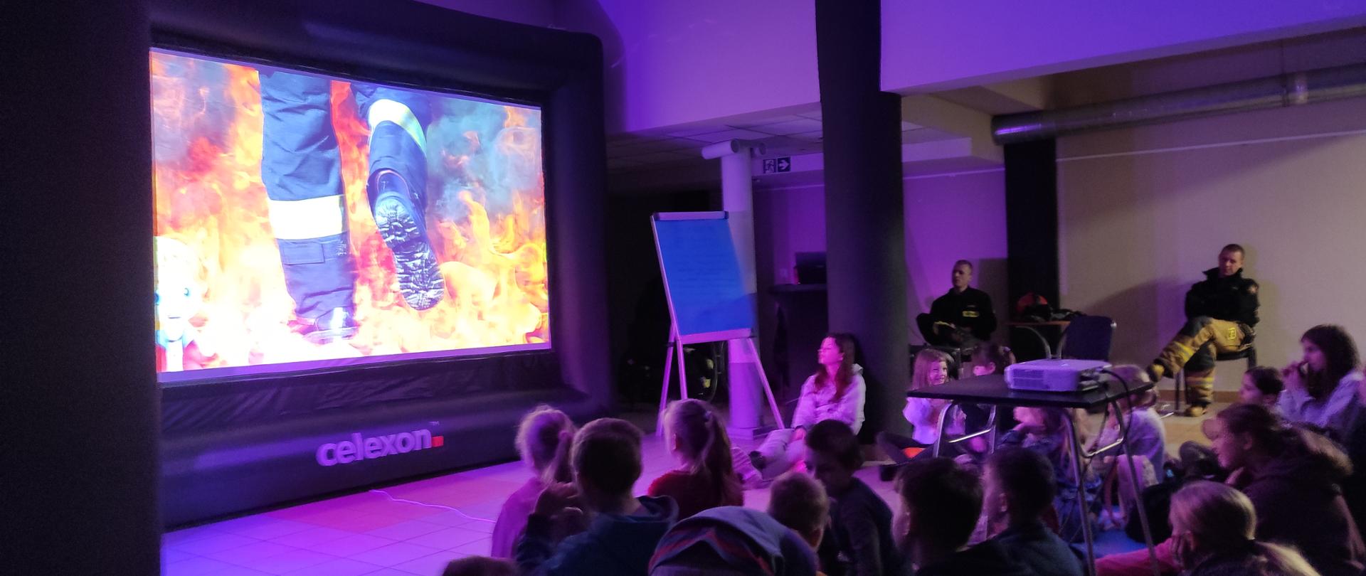 Wnętrze sali, zaciemnione światło, poświata fioletowego koloru. Na dole zdjęcia, dzieci siedzą na podłodze oglądają film o strażakach puszczany z rzutnika. 