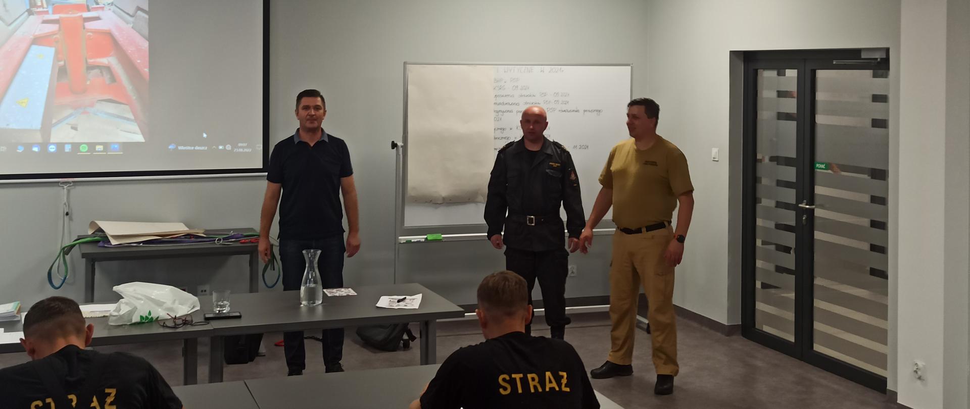 Zdjęcie przedstawia strażaków podczas szkolenia. Przed strażakami stoją od lewej: prelegent, przedstawiciel Ośrodka Szkolenia przy Komendzie Wojewódzkiej PSP oraz dowódca Jednostki Ratowniczo-Gaśniczej numer 1 w Kielcach.