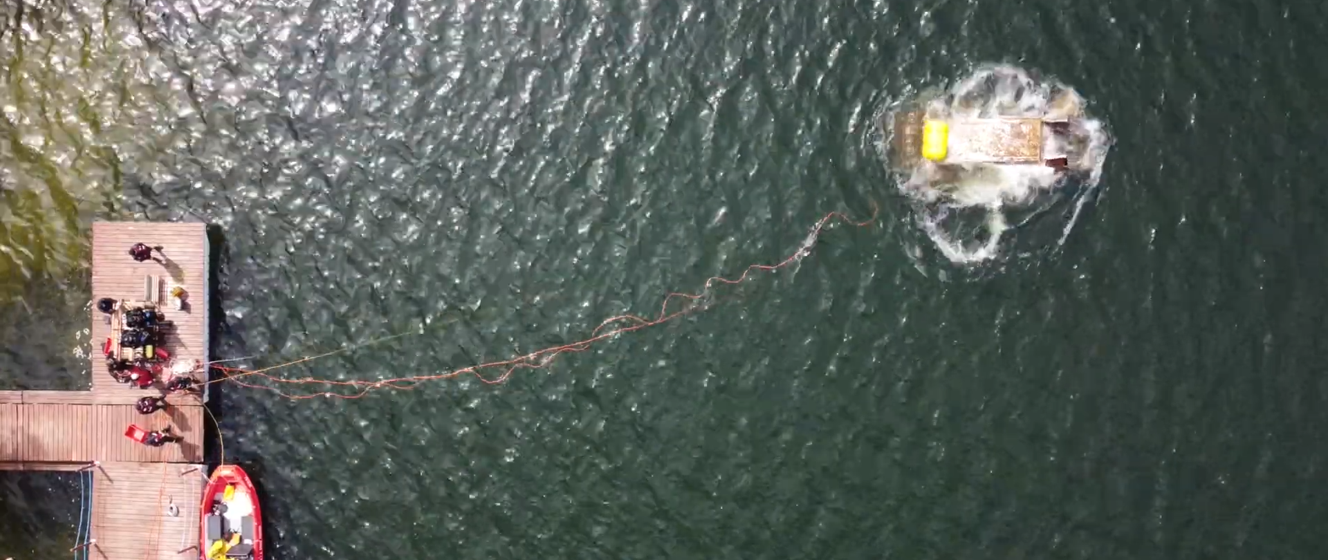 Ujęcie z lotu ptaka przedstawiające moment wynurzenia się wraku samochodu dostawczego. Do auta przymocowany jest żółty balon wypornościowy oraz liny i przewody powietrzne ciągnące się do pomostu. Na pomoście można zauważyć grupkę ludzi oraz różny specjalistyczny sprzęt ratownictwa wodno-nurkowego. Przy pomości stoi zacumowana łódź ratownicza.
