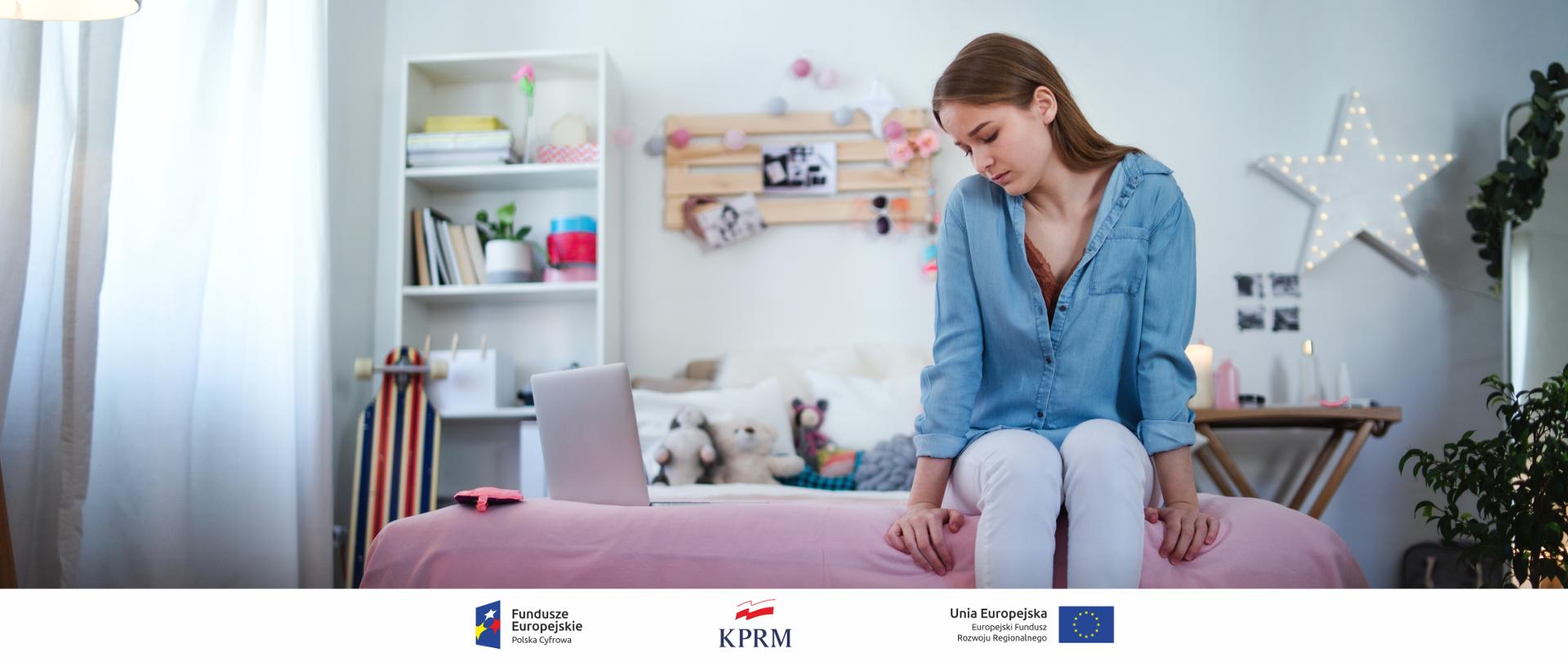 Smutna nastolatka siedzi na łóżku w swoim pokoju, spogląda w stronę laptopa.