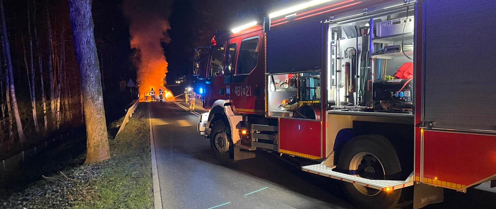 Zdjęcie przedstawia pojazd w płomieniach i strażaków gaszących pożar 
