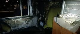 Wnętrze mieszkania po pożarze. Pod oknem spalone łóżko, ściany oraz okno okopcone.