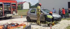 Strażacy wkładający klin pod koła samochodu wykorzystanego do pozoracji, po lewej stronie samochód pożarniczy oraz sprzęt hyddauliczny na macie. 