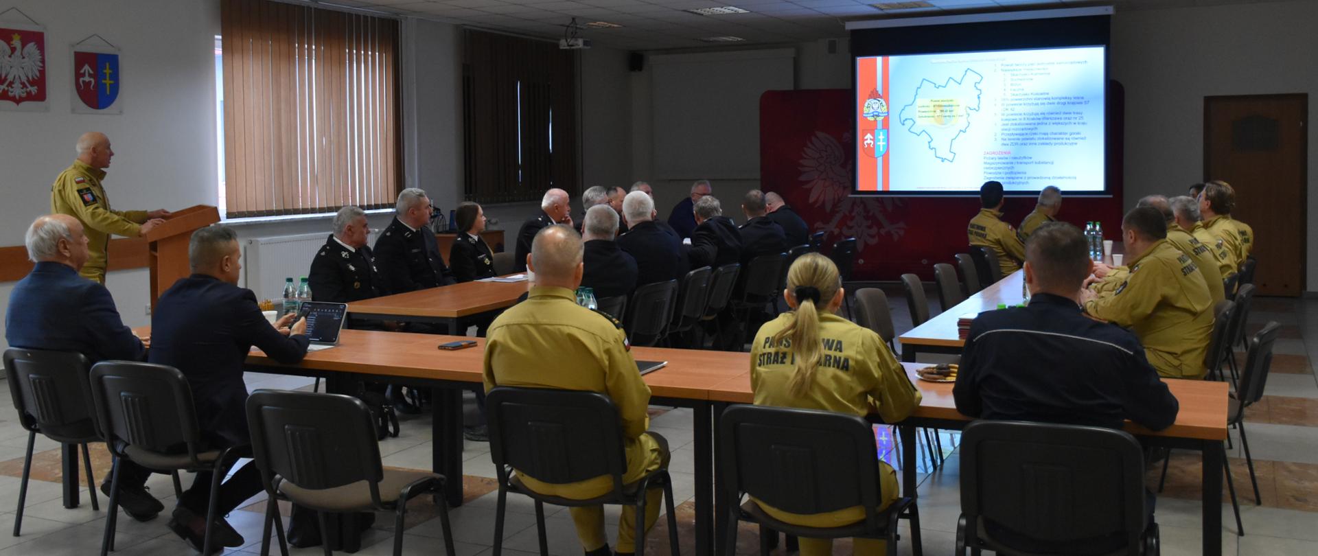 W dużej sali odpraw odbywa się prezentacja dla strażaków. Uczestnicy siedzą przy stołach, a na ich plecach widnieje napis „Państwowa Straż Pożarna”. Z przodu sali stoi mównica oraz duży ekran, na którym wyświetlana jest mapa i slajdy dotyczące działalności PSP. Obok ekranu wiszą dwa godła – polskie godło państwowe oraz emblemat PSP. Atmosfera jest formalna, typowa dla narady służbowe