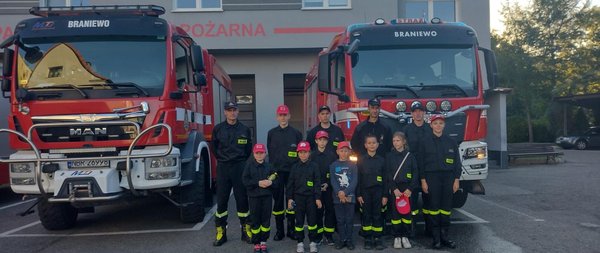 Na tle dwóch samochodów strażackich, stoją wspólnie dzieci z Młodzieżowej drużyny pożarniczej i strażacy. W oddali budynek strażnicy.
