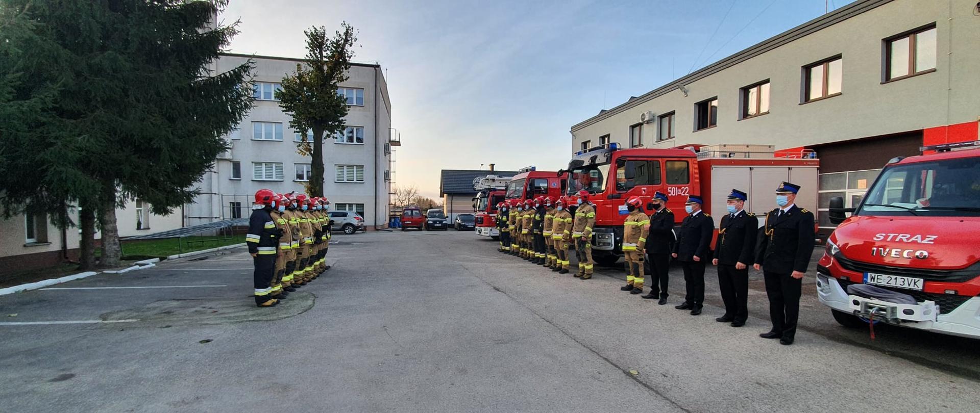 Zbiórka strażaków na placu wewnętrznym KP PSP Maków Mazowiecki. Dwie zmiany służbowe stoją naprzeciw siebie, ubrani w ubrania specjalne, czterech z nich ma ubrania wyjściowe. Po jednej stronie stoją przed budynkiem urzędu miasta, po drugiej stronie przed budynkiem komendy i czerwonymi samochodami strażackimi.