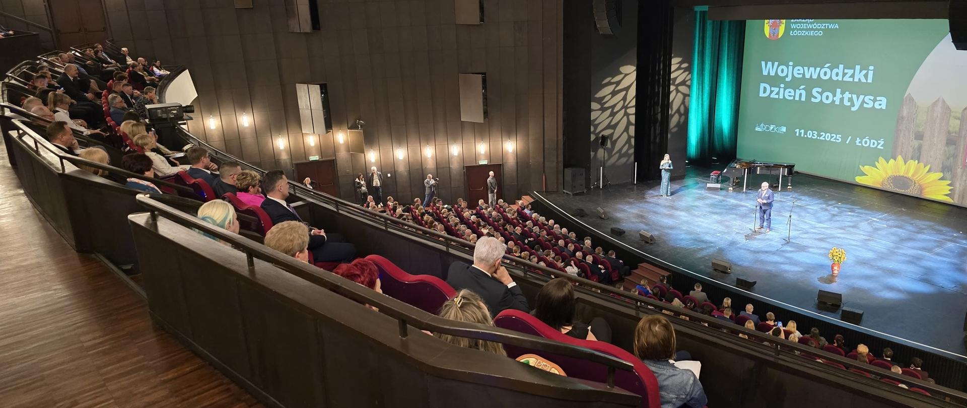 Widownia teatralna , na środku sceny przy mikrofonie stoi mężczyzna, a po lewej kobieta, na ekranie napis Wojewódzki Dzień Sołtysa.