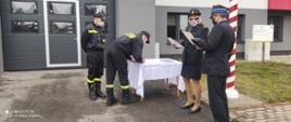 Nowo przyjęty strażak na białym stoliku podpisuje akt ślubowania obok niego stoi drugi strażak, przy stoliku z drugiej strony stoją komendant oraz asystująca. W tle budynek komendy. 