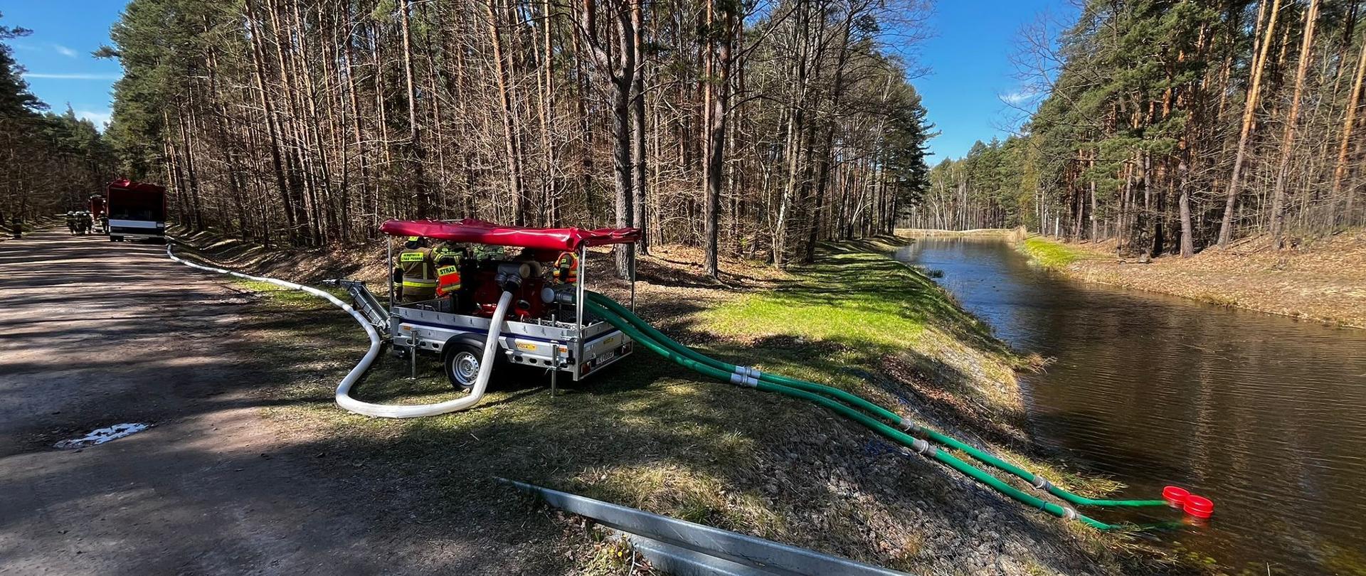 Fotografia przedstawia przyczepę z motopompami stojącą na brzegu cieku wodnego: przez 2 węże ssawne zasysana jest woda i podawana przez pompę w miejsce pożaru.