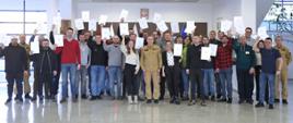 Pamiątkowe zdjęcie grupowe uczestników szkolenia inspektorów ochrony przeciwpożarowej (28 osób) w towarzystwie Zastępcy Komendanta Centralnej Szkoły PSP, dwóch funkcjonariuszy CS PSP oraz kobiety. Uczestnicy szkolenia trzymają radośnie w górze otrzymaną biała teczkę z zaświadczeniem o ukończeniu szkolenia