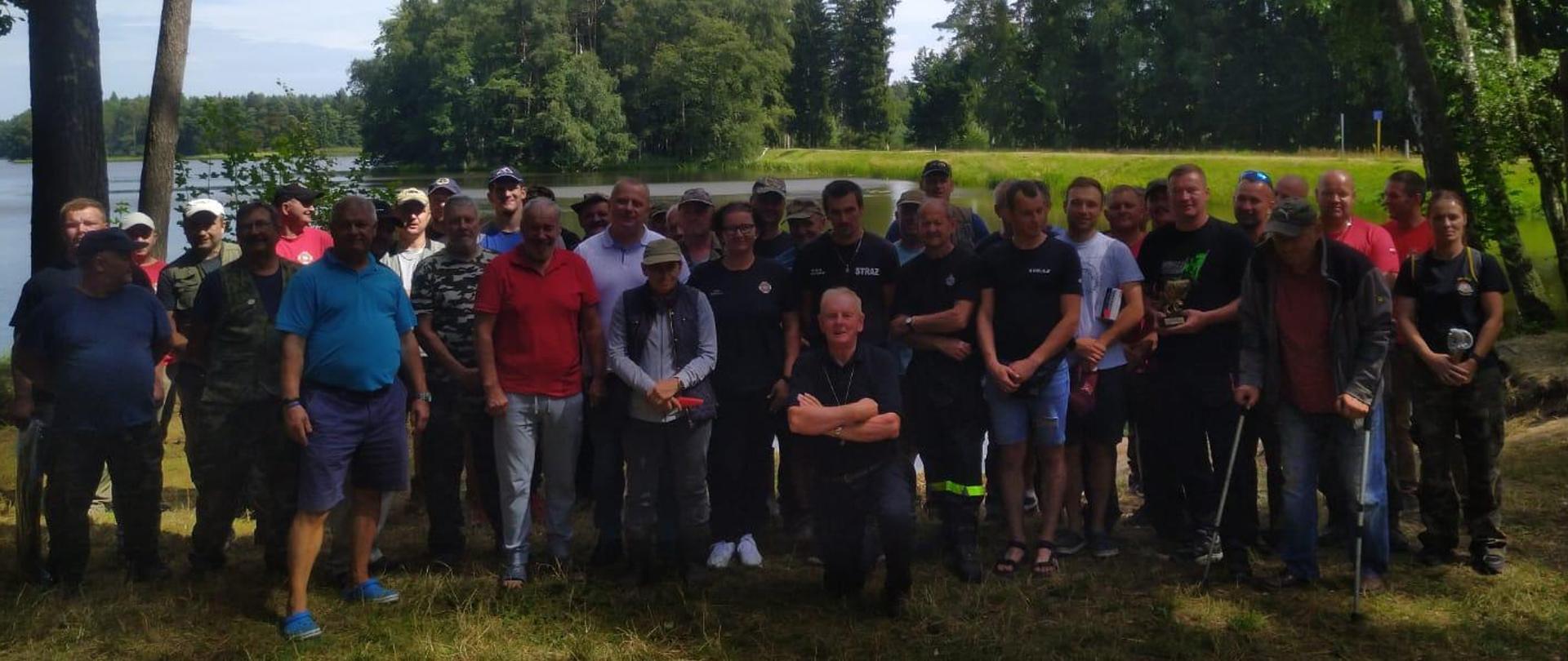 Zdjęcie grupowe strażaków, uczestników zawodów wędkarskich. W tle widok na wodę zbiornika rzeki Gwdy. 