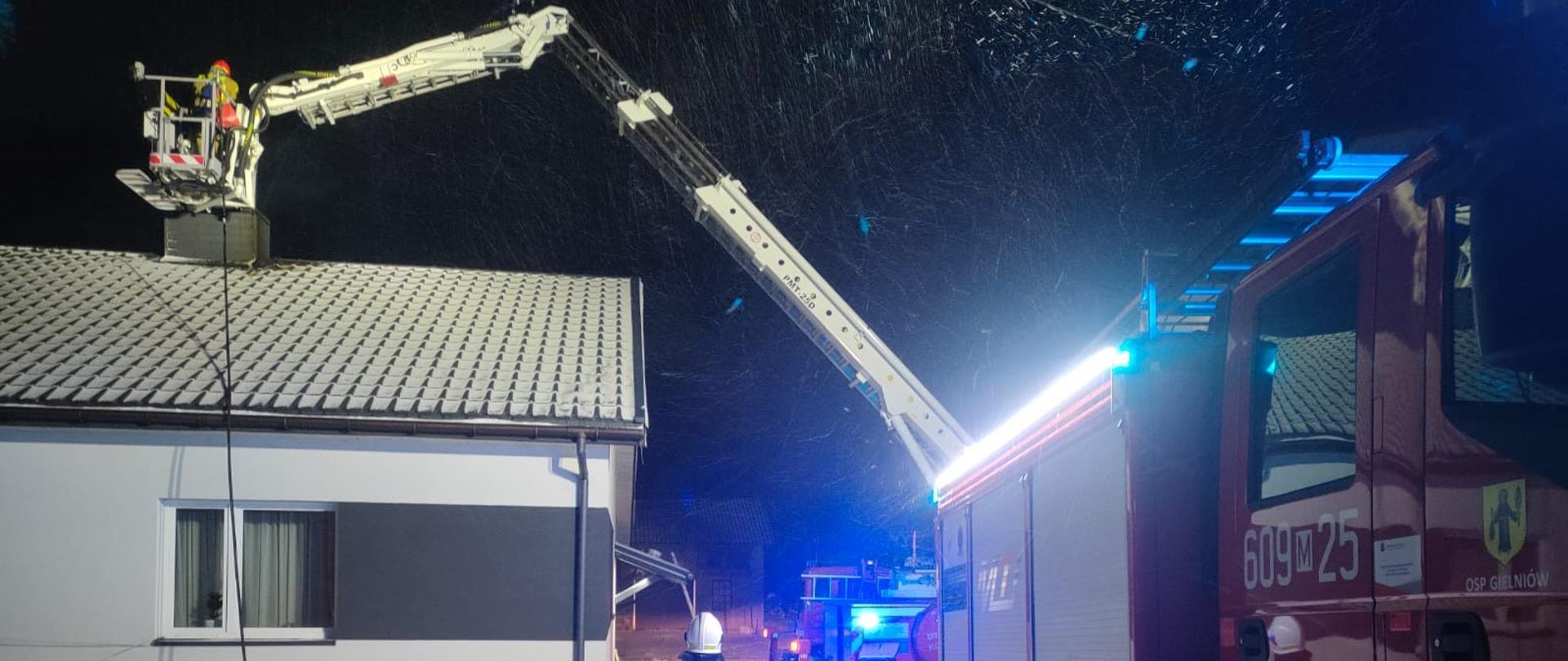 Zdjęcie przedstawia scenę nocnej akcji straży pożarnej w zimowej scenerii. Na pierwszym planie po prawej stronie widoczna jest część dużego czerwonego wozu strażackiego z niebieskimi światłami sygnalizacyjnymi, a na jego boku widnieje numer "609 25". Od drugiego wozu odchodzi długa, biała drabina hydrauliczna, która unosi się w lewo i do góry, kierując się nad dach białego domu z ciemnym dachem, pokrytego śniegiem. Na końcu drabiny, w koszu, widać strażak pracującego na wysokości. Na pierwszym planie, w centrum, stoi strażak w ciemnym kombinezonie odblaskowym z napisem "STRAŻ" na plecach, zwrócony tyłem do widza, obserwujący sytuację. Teren wokół jest zaśnieżony, a z nieba spadają drobne płatki śniegu, widoczne jako jasne punkty w blasku świateł. W tle, za domem, widać ciemne drzewa i inne światła. Cała scena jest oświetlona przez światła pojazdów i lampy, co tworzy silne kontrasty i wskazuje na dynamiczne działania ratownicze w trudnych warunkach pogodowych.