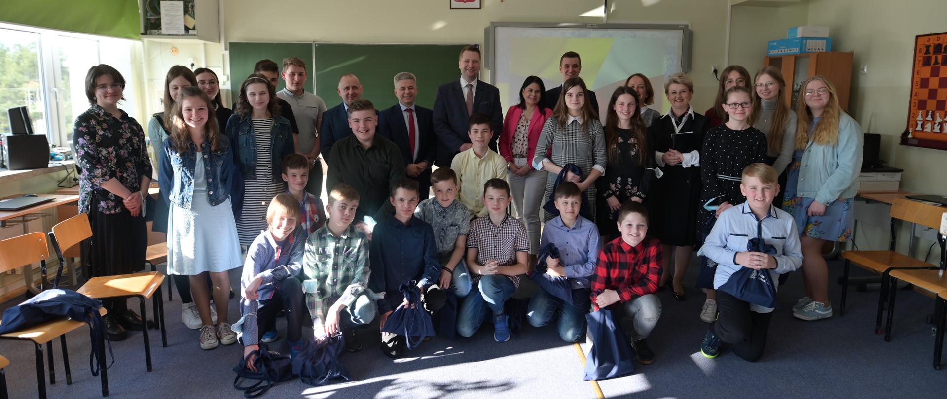 Grupa dzieci stojących i kucających w sali. W tle tablica i tablica multimedialna. Po obokach stoją krzesła. W otoczeniu uczniów minister Przemysław Czarnek. 
