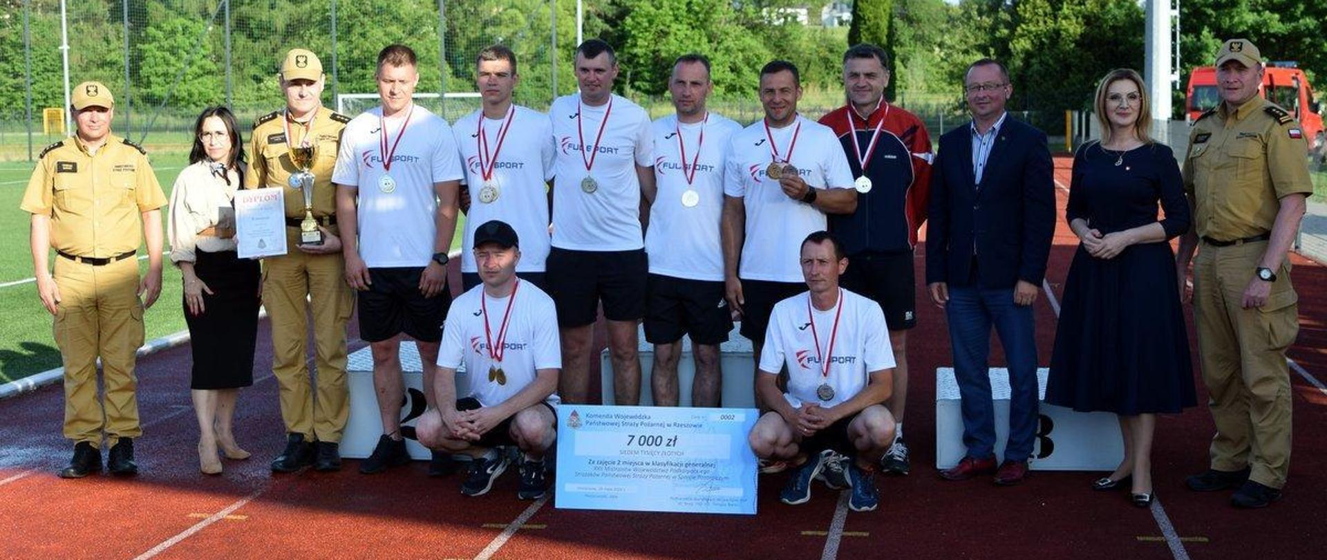Kolorowa fotografia wykonana w pogodny dzień na zewnątrz. Przedstawia strażaków sportowców po wręczeniu medali i nagród w sporcie pożarniczym . Z boku komendant i osoby wręczające.