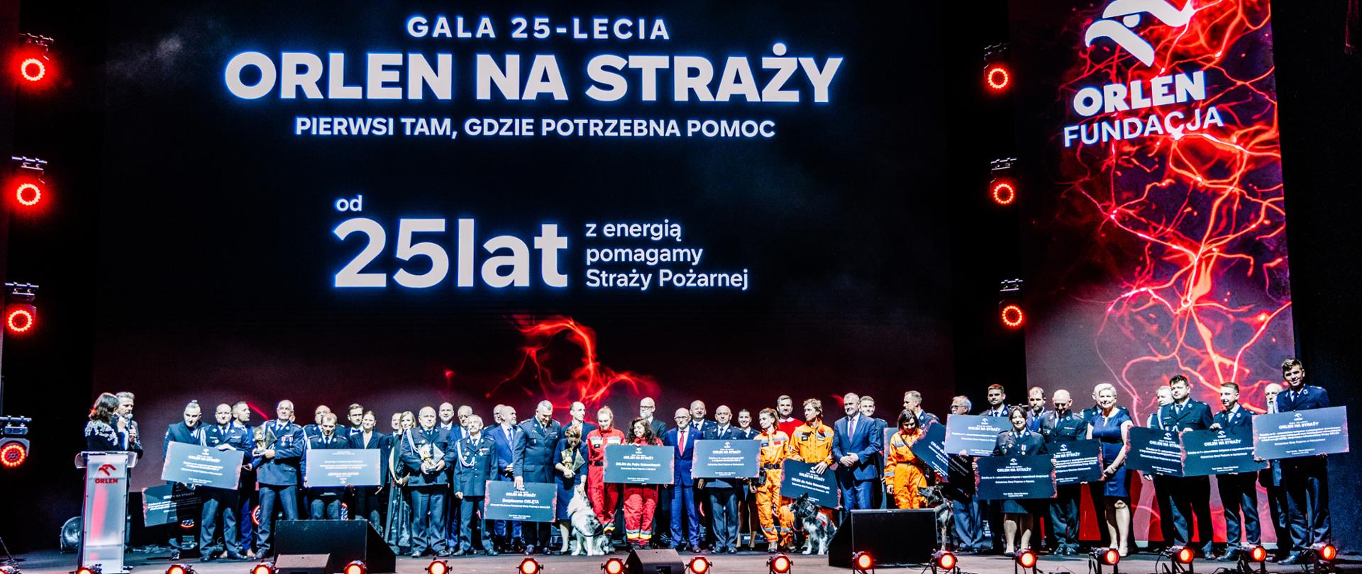 Uczestnicy i laureaci gali „ORLEN na Straży” zgromadzeni na scenie z pamiątkowymi tablicami. W tle ekran z napisem „Od 25 lat z energią pomagamy Straży Pożarnej”. 