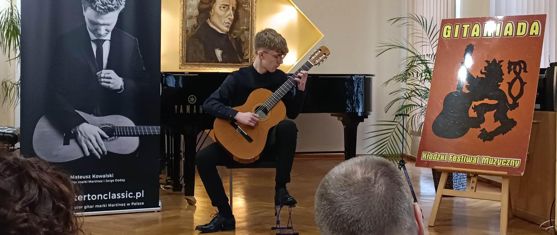 Nikodem Klatka - uczeń klasy gitary p. Przemysława Kolarskiego - grający na gitarze. W tle fortepian, a nad nim podświetlony portret Fryderyka Chopina. Z prawej strony na sztaludze brązowy plakat GITARIADY - Kłodzkiego Festiwalu Muzycznego (napisy w kolorze żółtym z czarnym obramowaniem i logo przedstawiające lwa z gitarą w kolorze czarnym). Z lewej banner reklamowy gitar marki Martinez w kolorze biało-czarnym. 
